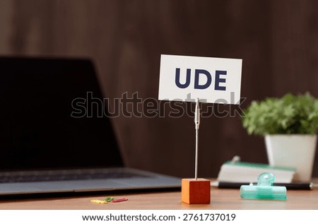 Eye-catching image of UDE text on card. The word stands for Undesirable Effect. Shown with laptop, clips, pen.