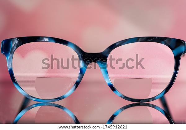 Eyeglasses Glasses with Bifocals and Black and\
blue Frame smudged view against a blurry pink  background with\
white hearts. Blurry Vision\
Concept