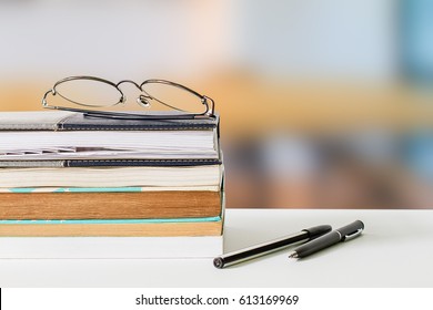 Eyeglasses on books and pens on white table on blurred library background