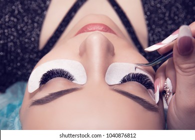 Eyelash extension process. Closeup portrait of young girl, woman with long and thick eyelashes, eyes closed and hand of a cosmetologist adding more eyelashes to her.