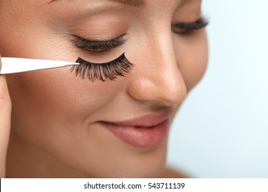 Eyelashes. Beautiful Woman Applying False Eyelashes With Tweezers. Closeup Of Young Female Model Face With Professional Facial Makeup, Smooth Skin And Long Black Thick Eye Lashes. High Resolution
