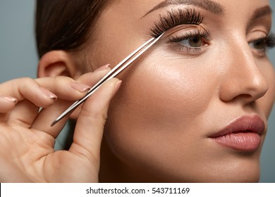 Eyelashes. Beautiful Woman Applying False Eyelashes With Tweezers. Closeup Of Young Female Model Face With Professional Facial Makeup, Smooth Skin And Long Black Thick Eye Lashes. High Resolution
