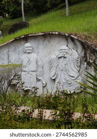 An eye-level, low-angle shot features a large rock carving on a grassy slope, depicting two smiling figures, likely historical or mythological, in detailed traditional robes.