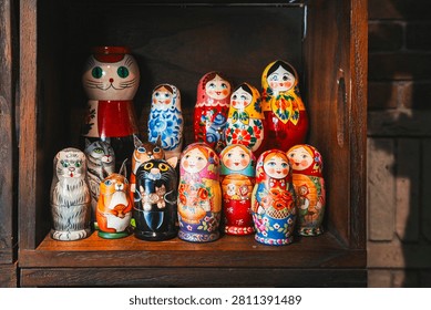 An eye-level, medium close-up shot capturing a colorful display of traditional Russian nesting dolls (matryoshka dolls) arranged inside a rustic wooden shelf or store window in the Motomachi shopping district of Yokohama, Japan. The collection features a mix of classic painted wooden female figurines wearing vibrant headscarves (babushkas) adorned with intricate floral patterns, alongside unique animal-themed nesting dolls shaped like cats, a squirrel holding an acorn, and owls. The dolls are neatly arranged on a dark wood shelf, with a warm brick wall texture visible on the right edge of the frame, creating a charming and nostalgic retail display under soft indoor lighting.