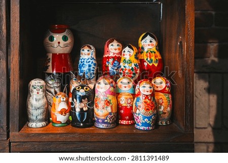 An eye-level, medium close-up shot capturing a colorful display of traditional Russian nesting dolls (matryoshka dolls) arranged inside a rustic wooden shelf or store window in the Motomachi shopping district of Yokohama, Japan. The collection features a mix of classic painted wooden female figurines wearing vibrant headscarves (babushkas) adorned with intricate floral patterns, alongside unique animal-themed nesting dolls shaped like cats, a squirrel holding an acorn, and owls. The dolls are neatly arranged on a dark wood shelf, with a warm brick wall texture visible on the right edge of the frame, creating a charming and nostalgic retail display under soft indoor lighting.