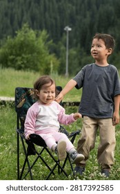 eye-level medium shot of two young children sitting next to each other in dark blue folding camp chairs with a colorful tropical leaf pattern. On the left, a young boy of Central Asian ethnicity is dressed in an oversized dark grey t-shirt and loose beige trousers, sitting calmly and looking toward his companion. On the right, a smaller toddler girl with fine brown hair is wearing a pink and white raglan long-sleeve top, light pink leggings, socks, and sneakers. She leans slightly toward the boy, gesturing or talking to him. They are positioned outdoors on a green grass lawn or meadow with a soft-focus background of lush green trees and a forested hill, capturing a candid, peaceful moment of childhood friendship and outdoor recreation.