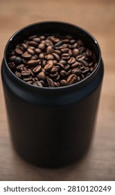 An eye-level, selective focus shot of aromatic medium-dark roasted coffee beans inside a modern matte black storage canister. A black plastic measuring scoop sits inside the jar, against a clean white surface and a soft, blurred background.