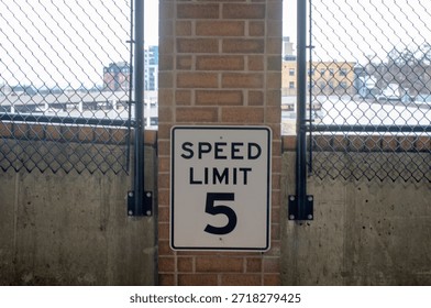 An eye-level shot of a rectangular white regulatory sign with the text "SPEED LIMIT 5" mounted on a red brick pillar inside an urban parking garage. The sign is positioned next to a black metal security fence, representing strict safety protocols and traffic management within a facility. The lighting is natural and bright, highlighting the contrast between the authoritative signage and the textured brickwork. This image conveys themes of property safety, facility management rules, and urban navigation. It is an ideal visual for content regarding parking garage security, liability insurance, and building management safety standards. The direct and clear composition makes the message the primary focal point, illustrating the regulatory side of shared residential or commercial parking decks. This photograph captures the daily reality of metropolitan building administration and the constant communication used to ensure safety in high-traffic utility zones.