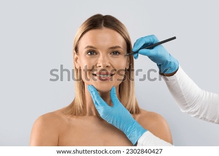 Eyelid surgery, laser blepharoplasty. Beautician hands drawing lines with pen on middle aged woman face, beautiful cheerful blonde lady have plastic surgery at clinic, copy space