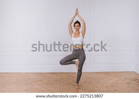 Eyes closed young brunette hispanic woman in sportswear standing in yoga balance pose at home. Mental health, healthy lifestyle. Pretty caucasian girl in meditation. Sport, hobbies, women.