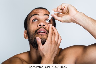 Eyes Feel Tired. Portrait Of Handsome Shirtless Young Black Man Using Eye Drops While Standing Against White Background
