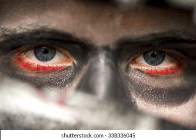 Eyes of a man wearing skull makeup with bloodshot rims and deep shadowed eye sockets looking at the camera over the blade of a knife, Halloween horror concept