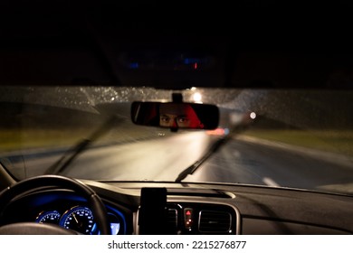 Eyes of young man are reflected in rearview mirror during night trip around city. Taxi driver or evening courier is driving along dark highway in passenger car with blue backlight of speedometer