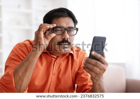 Eyesight Problems Concept. Mature Indian Man In Eyeglasses Looking At Smartphone Screen And Squinting, Trying To Read Message, Suffering From Astigmatism And Bad Vision, Closeup Shot, Copy Space