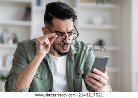 Eyesight Problems Concept. Young Arab Man In Eyeglasses Looking At Smartphone Screen And Frowning, Millennial Guy Trying To Read Message, Suffering From Astigmatism And Bad Vision, Closeup Shot
