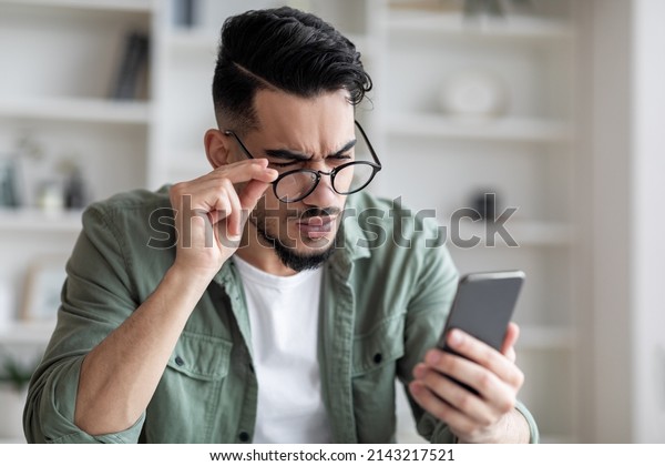 Eyesight Problems Concept. Young Arab Man In\
Eyeglasses Looking At Smartphone Screen And Frowning, Millennial\
Guy Trying To Read Message, Suffering From Astigmatism And Bad\
Vision, Closeup Shot