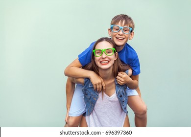  Eyewear concept. The gorgeous freckled brother and sister in casual t shirts wearing trendy glasses over background together. Brother climbed up on the back of a cute sister. Studio shot