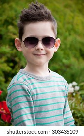 Eyewear Concept. Stylish Baby Boy With Dark Hair In Trendy Sunglasses  And In A Striped Polo-neck, The  Standing, In The Park With Green Grass. Hipster Style. Sunny Weather. Close Up. Outdoor Shot