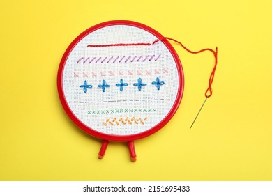 Fabric with colorful stitches in embroidery hoop and needle on yellow background, top view