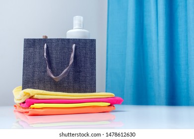 Fabric Freshener Spray, Fabric Softener In A Paper Bag Stands Next To A Stack Of Clean, Ironed Colored Laundry. Textile And Clothing Care Concept.