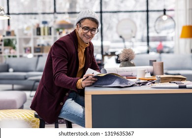 Fabric Samples Selection. Joyful Good-appealing Adult Fancy Designer In Stylish Knitted Hat, Glasses And Marsala Jacket Selecting Fabric Samples In Office