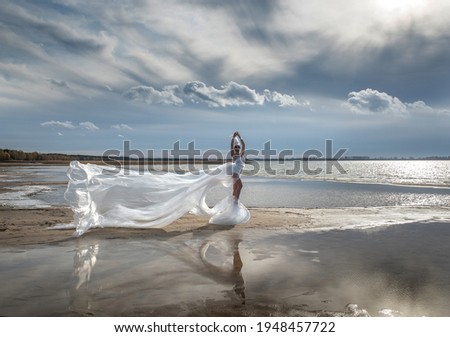 A fabulous girl in a long transparent dress fluttering in the wind and a high headdress on the shore of a reservoir.