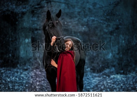 Fabulous portrait of a beautiful young lady with long brown hair, suite with red or burgundy cloak with her black horse in winter