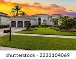 Facade of an elegant house with gray walls with white details, a red tiled roof, a front garden with abundant tropical plants, palm trees, short grass, sidewalk, driveway and garage