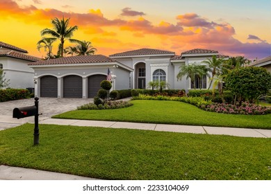 Facade of an elegant house with gray walls with white details, a red tiled roof, a front garden with abundant tropical plants, palm trees, short grass, sidewalk, driveway and garage - Shutterstock ID 2233104069