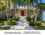 Facade of elegant and luxurious mansion, with palm trees in flower beds on the way to the main door, attractive red door combined with pastel and warm tones of the walls, driveway, short grass 