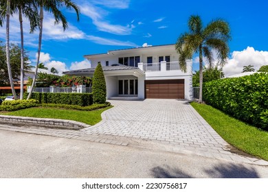 Facade of elegant modern house in the Riviera Isles neighborhood of Miami, privet wall, driveway to garage, balcony, black tiles, tropical vegetation and short grass, white walls, blue sky