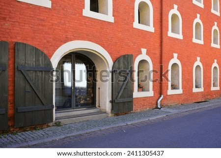 Facade of the historic building with small semicircular windows, antique doors with wooden sliding shutters,  red brick wall. Ancient architecture, old street. Torun, Poland, August 2023 