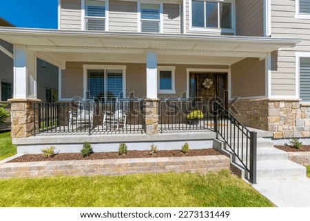Facade of a house with gray wood and stone siding and stone planters at the front of the porch. Porch of a house with railings and gray rocking chairs near the windows and front door with flowers.