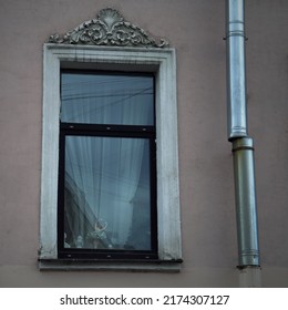 The Facade Of An Old 19th Century Residential Building In Historic Center Of St. Petersburg, Window Close Up - Beautiful Molding Above, Broken Drain Pipe On The Right, Curtains And Figurines Inside