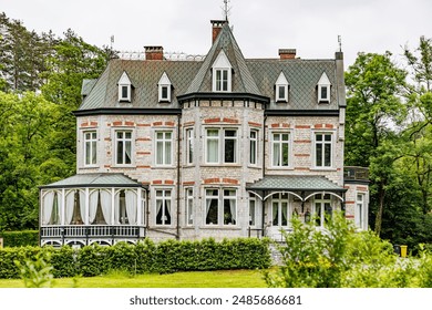 Facade of an old Victorian style house, white walls, gray gable roofs, multiple windows, closed veranda and garden, green leafy trees in background, cloudy day in Ferrieres, Belgium - Powered by Shutterstock