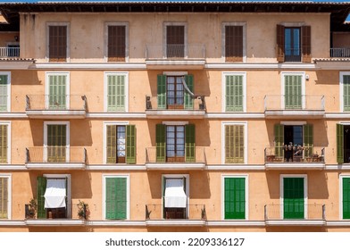 The Facade Of A Residential Building In Mallorca, Spain With Balconies And French Doors Partially Closed With Shutters In The Midday Sun