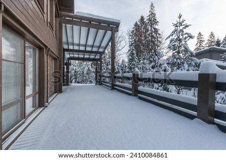 The facade of a wooden house with a spacious terrace against the background of snow-covered fir trees on a clear winter day.