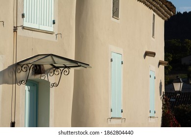 Facades of Provencal houses. Sand blond colour, with borders around the windows. Light green shutters. A black steel awning above the door.