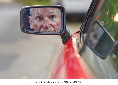 Face Of An Attractive Male Senior Is Reflected In The Rearview Mirror On Driver's Side Of A Red Car