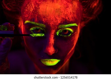 Face Of A Beautiful Young Sexy Girl With Ultraviolet Paint On Her Skin Painting Her Eyelashes With Brush In Bright Dye. Pretty Woman With Glowing Bodyart In Black Lamp Light