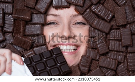 The face of a caucasian woman surrounded by chocolates. The girl eats a bar of chocolate.