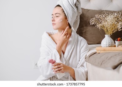 Face Cream Woman In Bathrobe With A Towel On Her Head After Showering At Home. Applying Skin Cream On Skin.