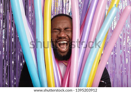 Face of dark skinned man through party modeling balloons, screams loudly, shows white perfect teeth, prepares for celebration special occasion. Unshaven young guy exclaims, has wide opened mouth