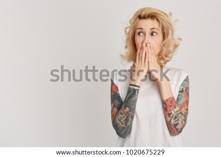 Face expression. Astonished and shocked young girl with tattoos over her arm looking aside and closed her mouth with hands. Emotions. Isolated on white wall. 
