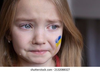 The Face Of A Frightened Girl, A Child Crying, Tears Flowing, Painted On Her Cheek In The Yellow-blue Colors Of The Ukrainian Flag, A Request For Help. Children Ask For Peace