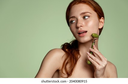 Face lift and anti-aging spa. Young redhead woman does gua-sha massage on face with jade stone roller, cosmetology treatment at home. Girl has glowing healthy skin and freckles, green background.
