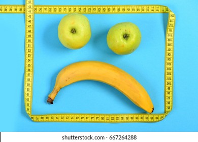 Face Made Of Fruit With Sad Expression: Banana And Apples Making Mouth And Eyes Framed With Yellow Flexible Ruler, Isolated On Cyan Blue Background. Concept Of Food Art And Minimalism