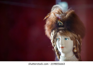 The Face Of An Old Retro Porcelain Doll In A Fur Hat On A Burgundy Background.