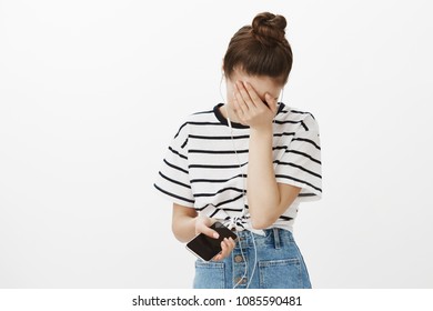 Face Palm From Stupid Song Lyrics. Portrait Of Devastated Upset Young Woman With Bun Hairstyle, Covering Face With Hand And Bending Head Down, Listening Sad Song In Earphones, Holding Smartphone