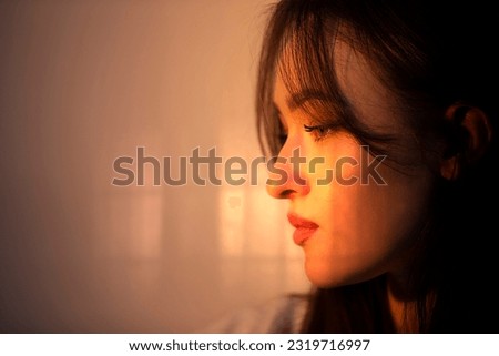 The face of a pensive girl in profile is illuminated by warm light from the window.
